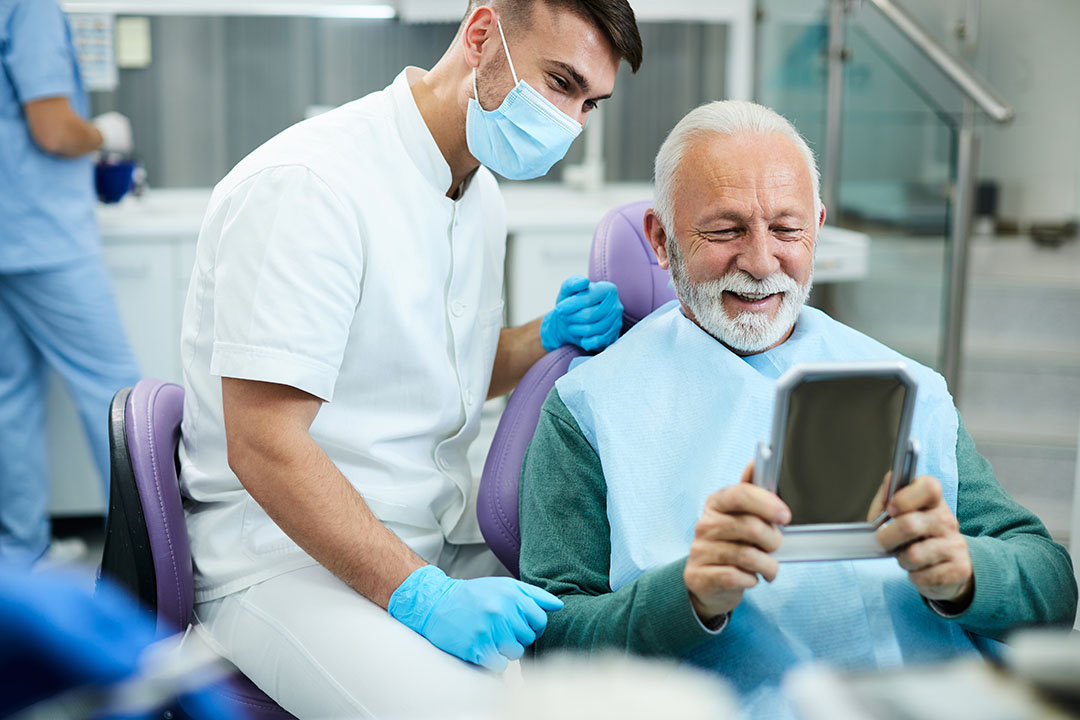 In the image, there is an elderly man sitting in a dental chair while a dentist attends to him, holding up a tablet with a blue screen.