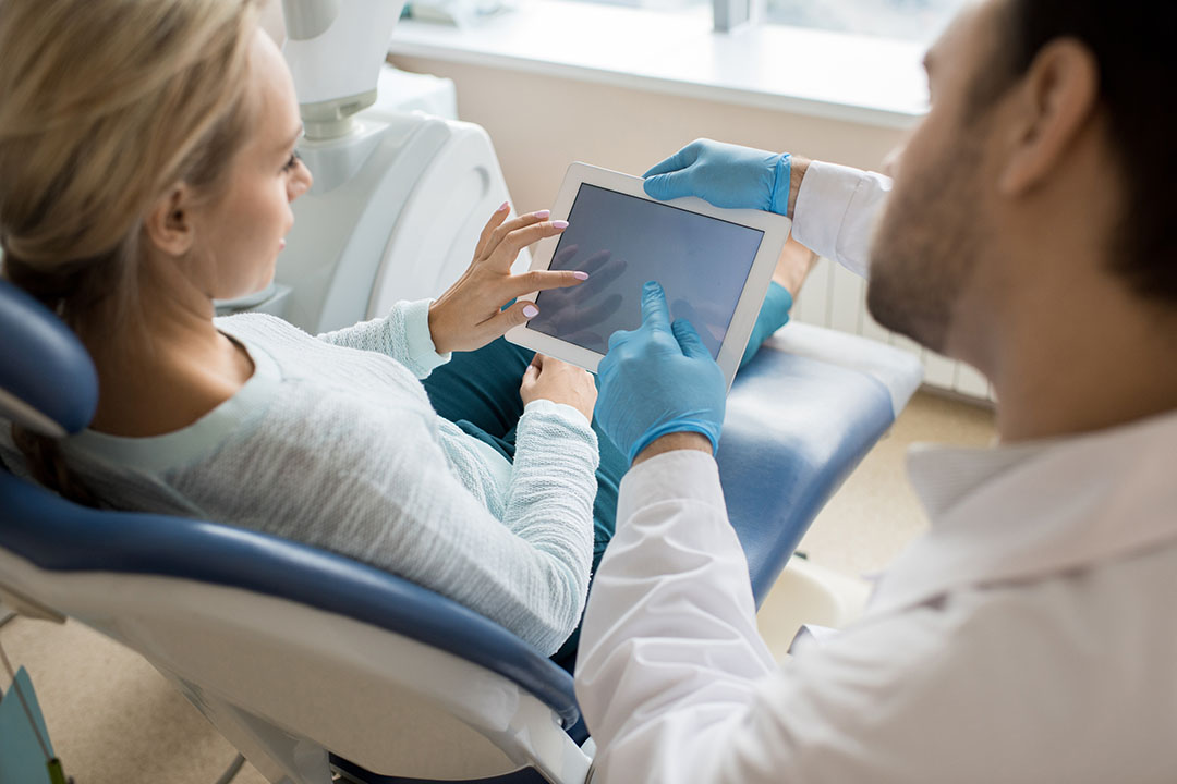 The image shows a dental professional assisting a seated patient with technology, likely during an examination or treatment planning session.