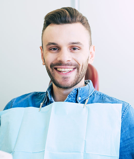 The image shows a man with a beard smiling at the camera while wearing a blue apron over a white shirt and sitting in front of a dental chair, indicating he works in dentistry.