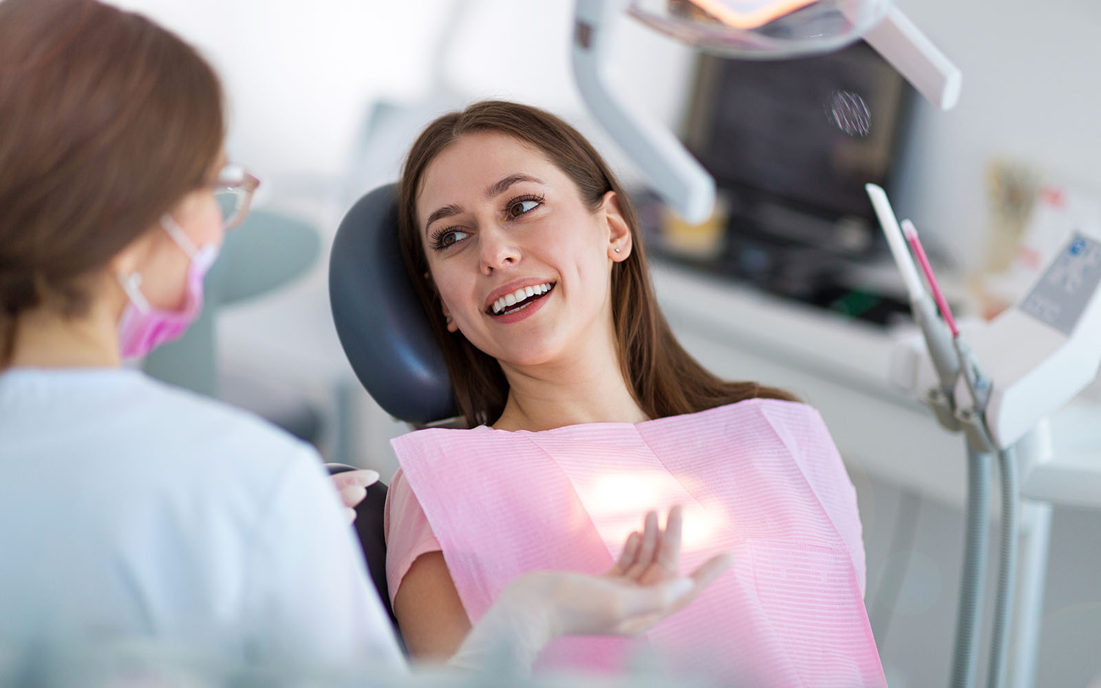 In the image, a woman is seated in an oral care chair, smiling at someone out of frame, while a dental professional stands nearby with a smile. They are both in a dental office setting, with equipment visible in the background.