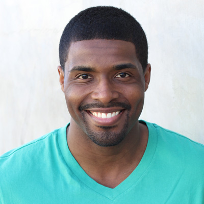 A man and woman are seated closely together, smiling at the camera, with the man wearing glasses and both dressed casually.