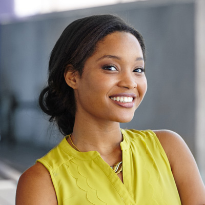 The image shows a smiling woman posing with her hands behind her back, wearing a yellow top and a necklace, against a blurred background that suggests an indoor setting.