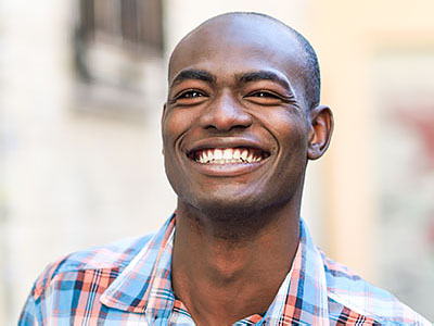 The image shows a smiling man with short hair, wearing a blue plaid shirt, standing outdoors against a blurred background.