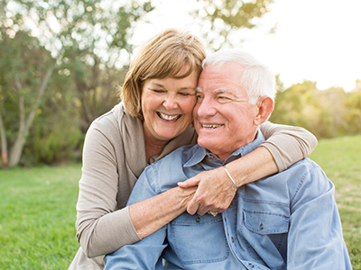 The image depicts an elderly couple embracing each other outdoors during daylight hours.