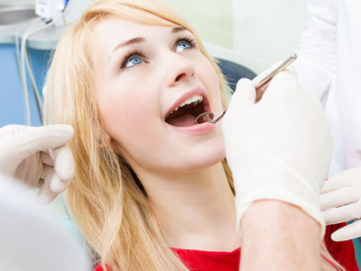 The image shows a woman sitting in a dental chair with her mouth open wide, receiving dental care from a professional wearing gloves and a face mask, who appears to be performing a procedure on her teeth.
