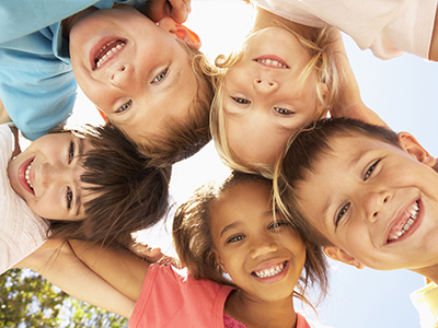 The image shows a group of children with different hair colors and styles smiling at the camera, with the sun shining brightly behind them.
