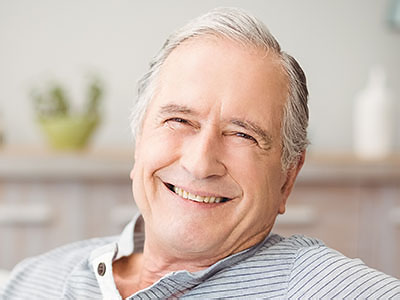 The image shows a smiling older man with gray hair, wearing a dark shirt, sitting comfortably with his arms crossed in front of him.